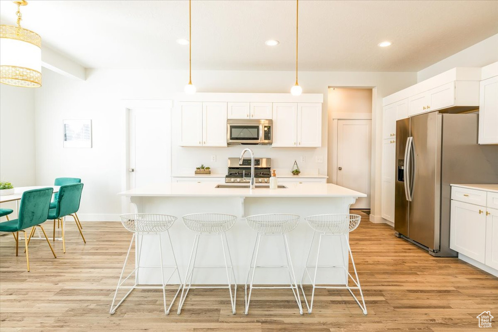 Kitchen featuring decorative light fixtures, light hardwood / wood-style flooring, stainless steel appliances, an island with sink, and white cabinets