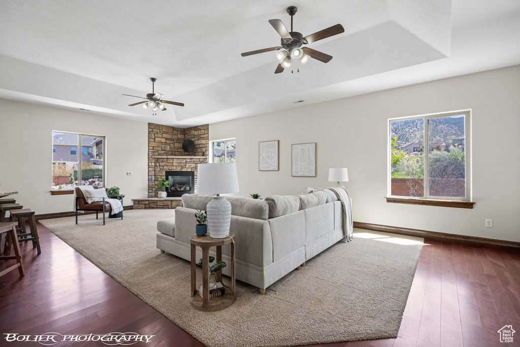 Living room featuring a stone fireplace, ceiling fan, a raised ceiling, and hardwood / wood-style flooring