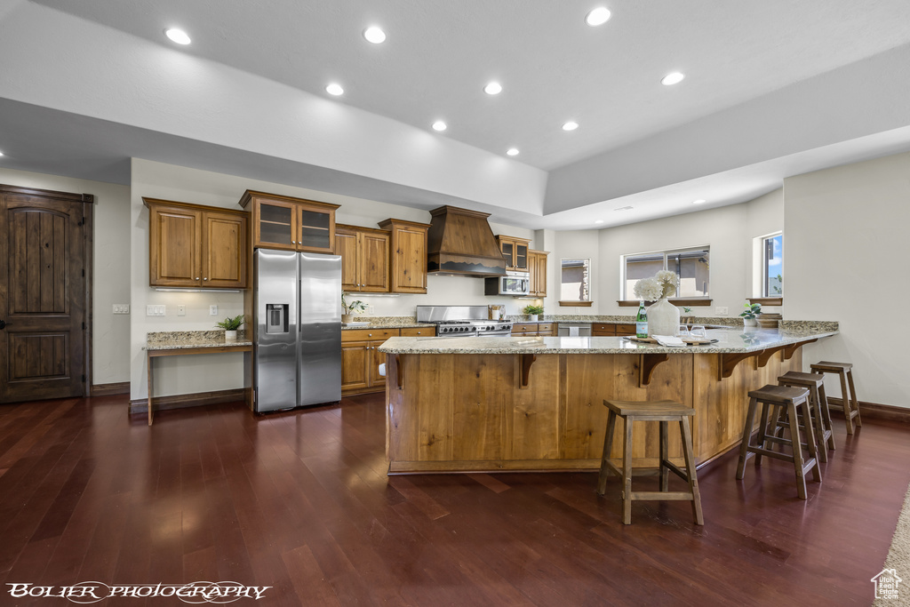 Kitchen featuring light stone counters, custom range hood, stainless steel appliances, and dark wood-type flooring