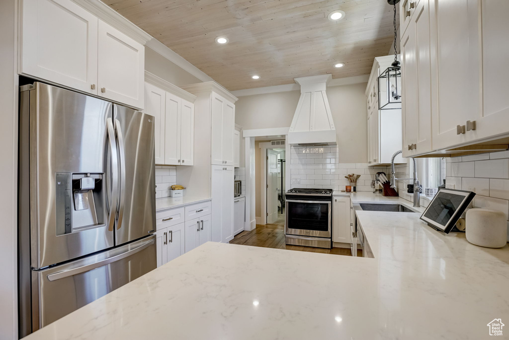Kitchen featuring custom exhaust hood, white cabinetry, appliances with stainless steel finishes, light stone counters, and tasteful backsplash