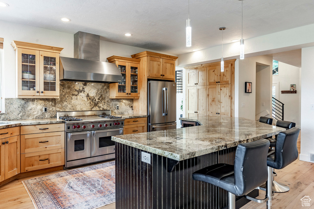 Kitchen featuring appliances with stainless steel finishes, light hardwood / wood-style flooring, backsplash, wall chimney exhaust hood, and a breakfast bar