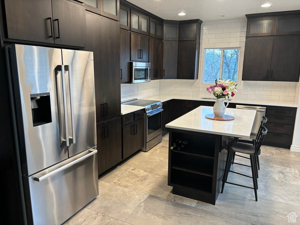 Kitchen with dark brown cabinetry, appliances with stainless steel finishes, a kitchen island, a kitchen bar, and tasteful backsplash