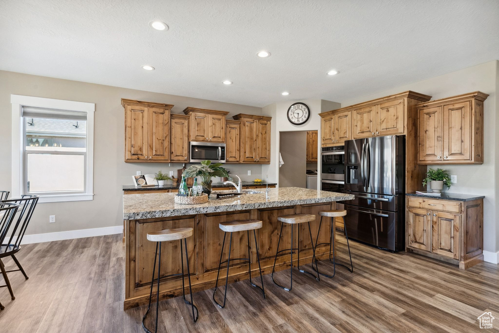 Kitchen with light stone counters, stainless steel appliances, a kitchen island with sink, wood-type flooring, and a breakfast bar