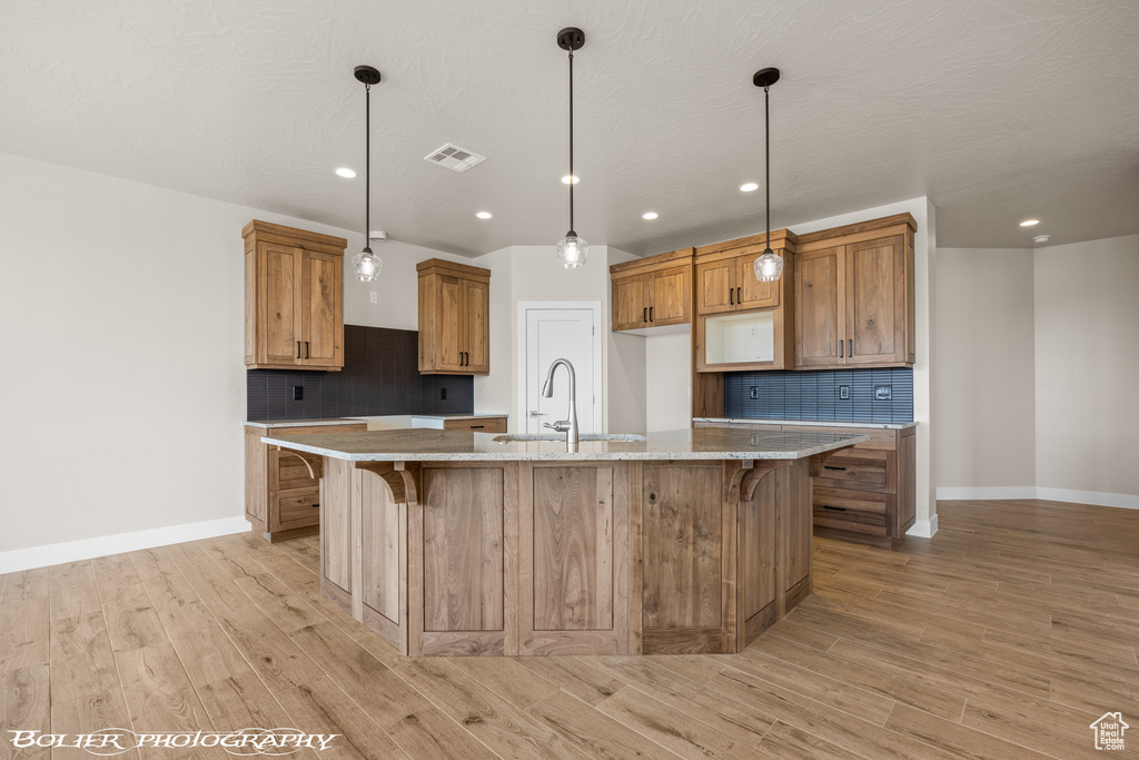 Kitchen with sink, light hardwood / wood-style floors, backsplash, an island with sink, and pendant lighting