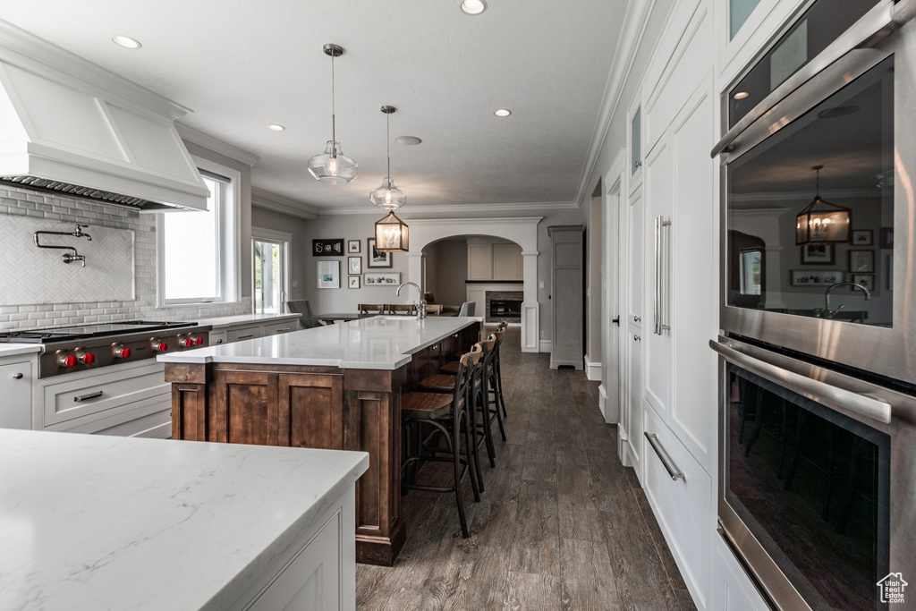Kitchen with backsplash, dark wood-type flooring, decorative light fixtures, a center island with sink, and premium range hood