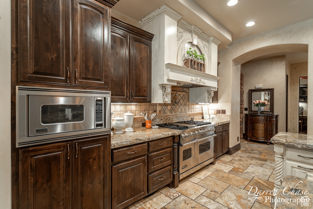 Kitchen featuring dark brown cabinets, appliances with stainless steel finishes, backsplash, light stone counters, and light tile floors