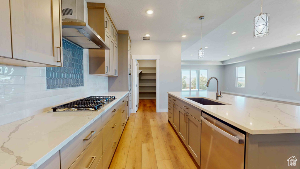 Kitchen featuring appliances with stainless steel finishes, a center island with sink, wall chimney range hood, sink, and light wood-type flooring
