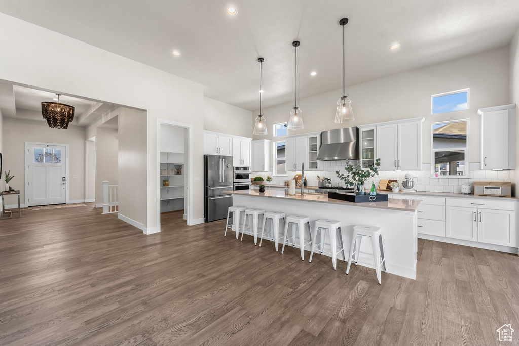 Kitchen with wall chimney exhaust hood, white cabinets, hardwood / wood-style flooring, tasteful backsplash, and an island with sink