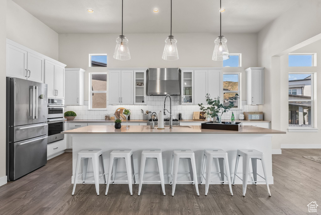 Kitchen featuring wood-type flooring, backsplash, wall chimney range hood, stainless steel appliances, and an island with sink