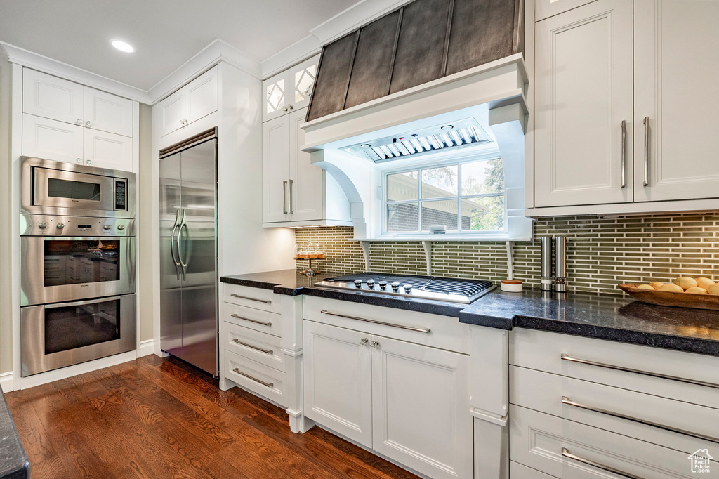 Kitchen featuring built in appliances, backsplash, dark wood-type flooring, white cabinetry, and dark stone countertops