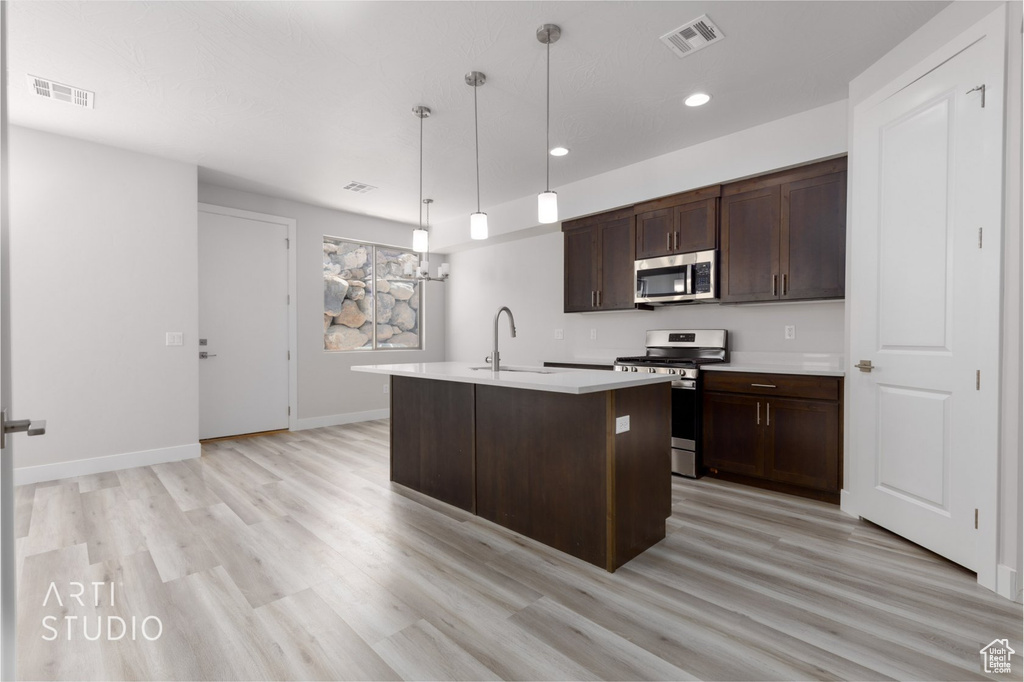 Kitchen featuring light wood-type flooring, dark brown cabinets, stainless steel appliances, an island with sink, and pendant lighting