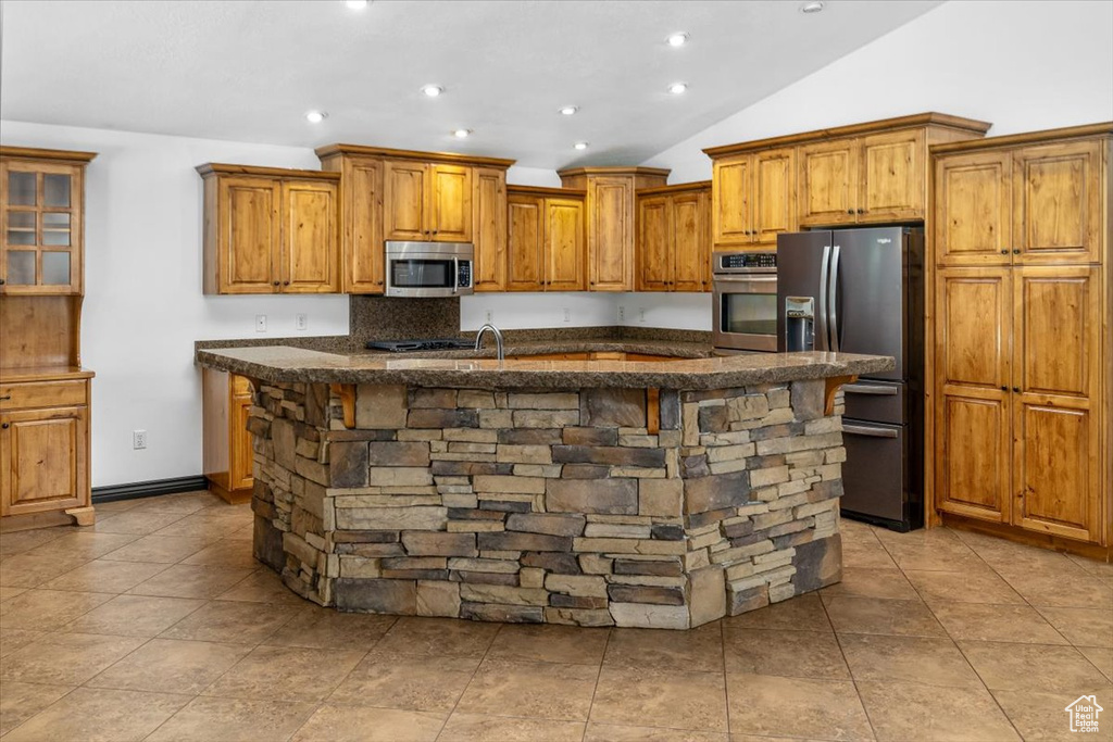 Kitchen featuring vaulted ceiling, appliances with stainless steel finishes, an island with sink, and light tile flooring