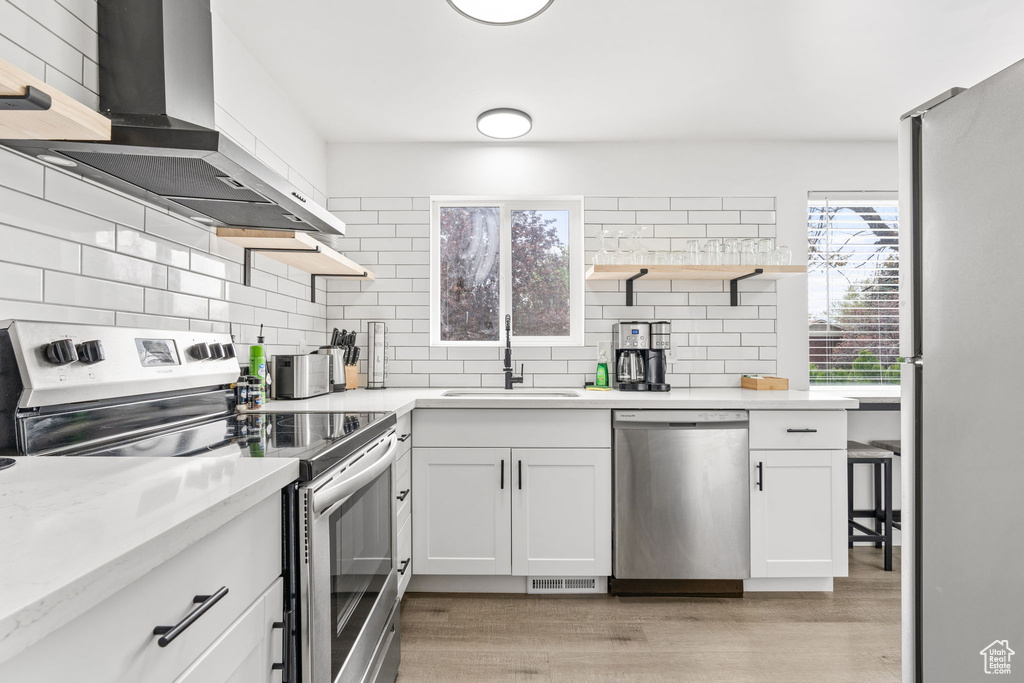 Kitchen featuring appliances with stainless steel finishes, white cabinets, light hardwood / wood-style floors, wall chimney range hood, and tasteful backsplash