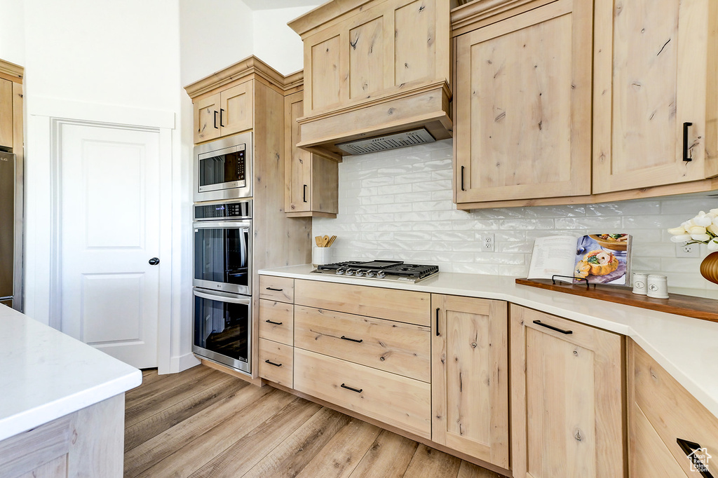 Kitchen with light wood-type flooring, tasteful backsplash, light brown cabinetry, premium range hood, and appliances with stainless steel finishes