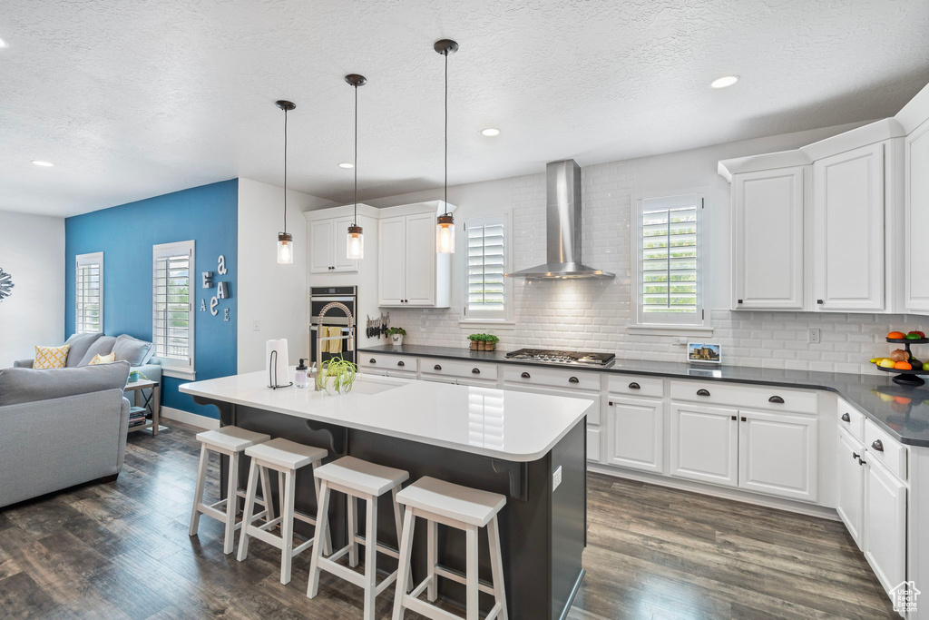 Kitchen with dark hardwood / wood-style floors, tasteful backsplash, an island with sink, wall chimney exhaust hood, and white cabinets