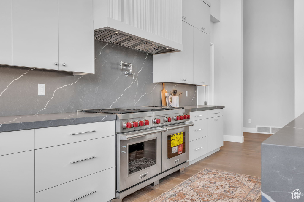 Kitchen featuring range with two ovens, light hardwood / wood-style flooring, tasteful backsplash, custom exhaust hood, and white cabinetry