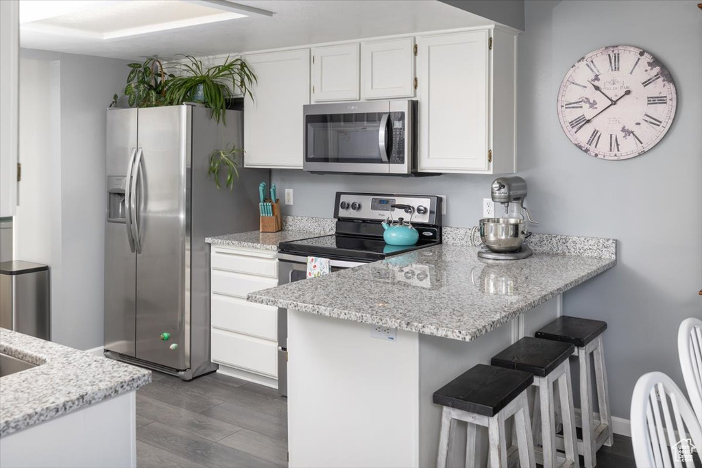 Kitchen with white cabinetry, stainless steel appliances, kitchen peninsula, light stone counters, and hardwood / wood-style floors