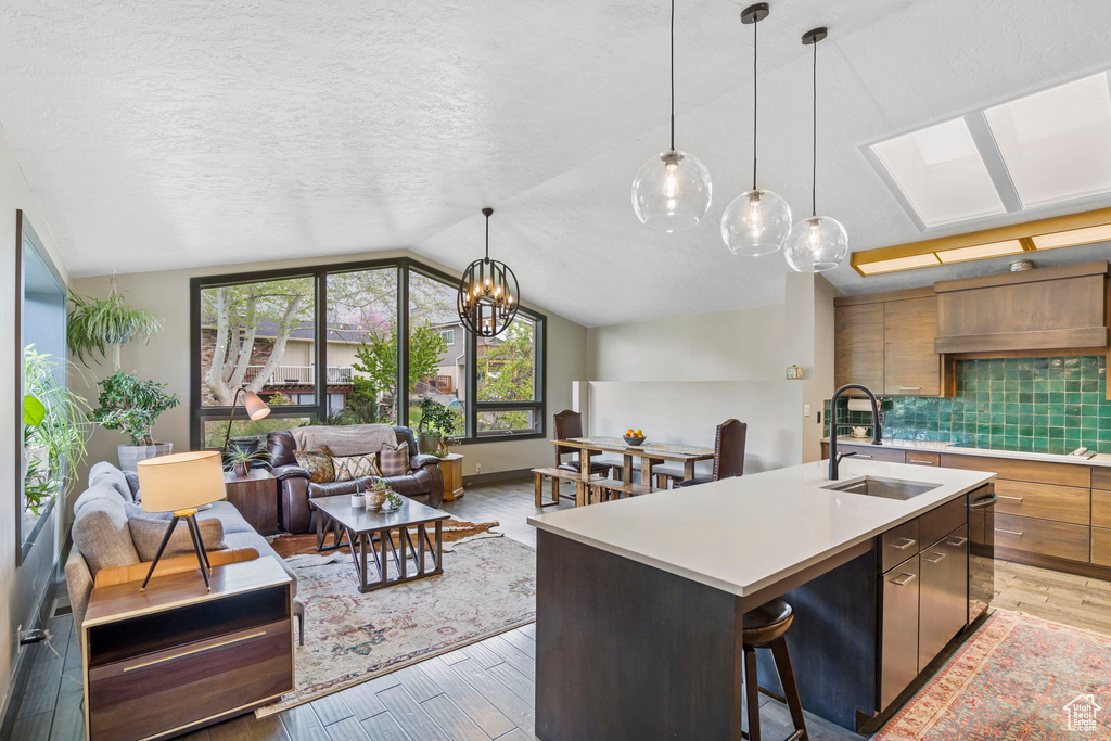 Kitchen with lofted ceiling with skylight, backsplash, sink, light hardwood / wood-style floors, and an island with sink