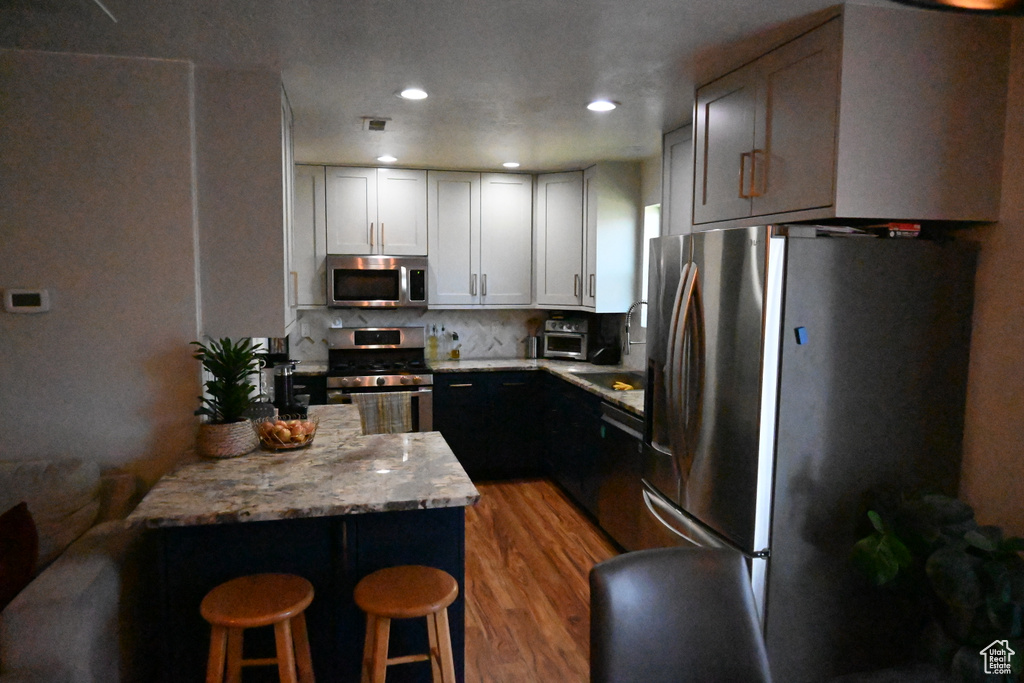 Kitchen with light wood-type flooring, appliances with stainless steel finishes, a breakfast bar area, light stone counters, and tasteful backsplash