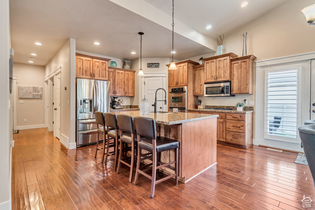 Kitchen featuring dark hardwood / wood-style floors, a center island with sink, decorative light fixtures, light stone counters, and appliances with stainless steel finishes