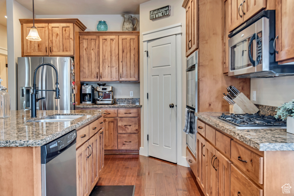 Kitchen featuring light stone countertops, appliances with stainless steel finishes, an island with sink, hardwood / wood-style flooring, and decorative light fixtures