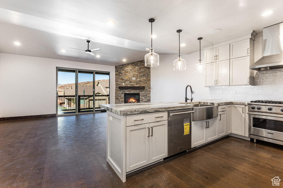 Kitchen featuring wall chimney range hood, dark hardwood / wood-style flooring, stainless steel appliances, ceiling fan, and a stone fireplace