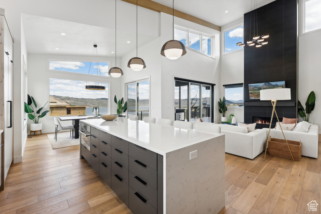 Kitchen with a towering ceiling, light hardwood / wood-style floors, hanging light fixtures, a tiled fireplace, and a kitchen island with sink