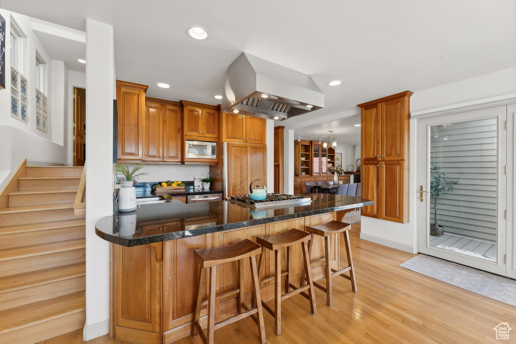 Kitchen featuring dark stone counters, light hardwood / wood-style flooring, island range hood, appliances with stainless steel finishes, and a breakfast bar area