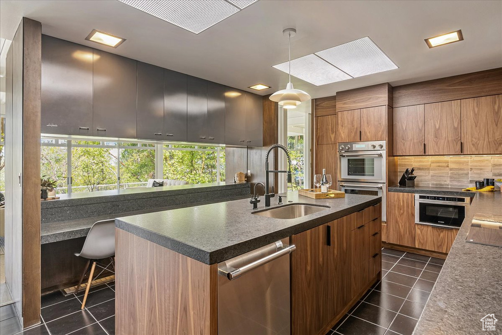 Kitchen featuring hanging light fixtures, a center island with sink, dark tile flooring, sink, and appliances with stainless steel finishes