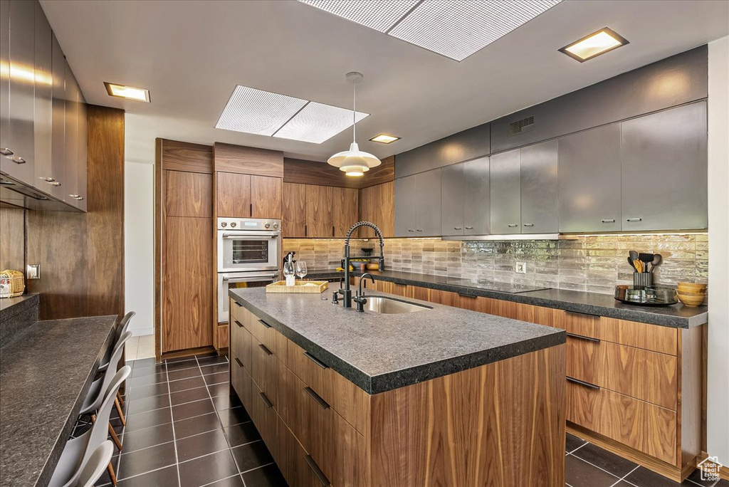 Kitchen featuring stainless steel double oven, a kitchen island with sink, decorative light fixtures, backsplash, and dark tile flooring