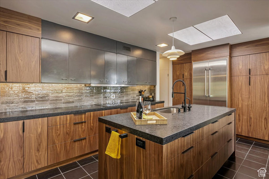 Kitchen with tasteful backsplash, an island with sink, stainless steel built in fridge, dark tile flooring, and sink