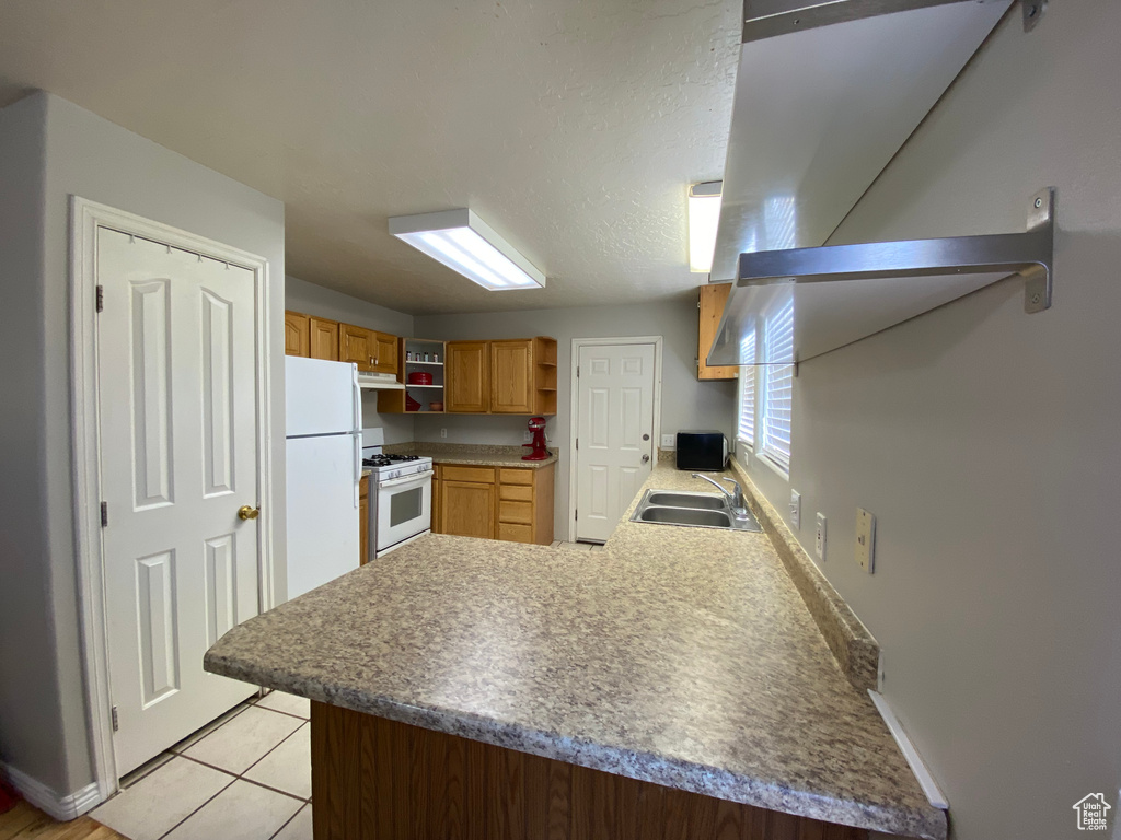 Kitchen with kitchen peninsula, white appliances, light tile flooring, wall chimney exhaust hood, and sink