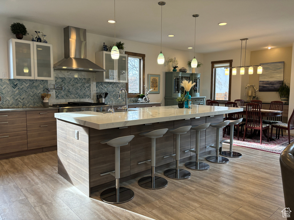 Kitchen featuring tasteful backsplash, an island with sink, hanging light fixtures, wall chimney exhaust hood, and sink