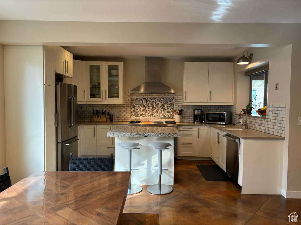 Kitchen featuring a kitchen island, appliances with stainless steel finishes, white cabinets, wall chimney exhaust hood, and tasteful backsplash