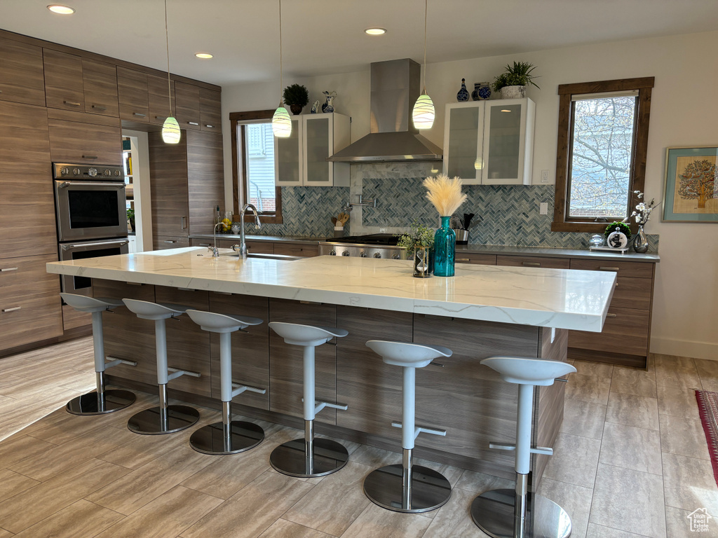 Kitchen with a center island with sink, light stone counters, backsplash, wall chimney range hood, and sink