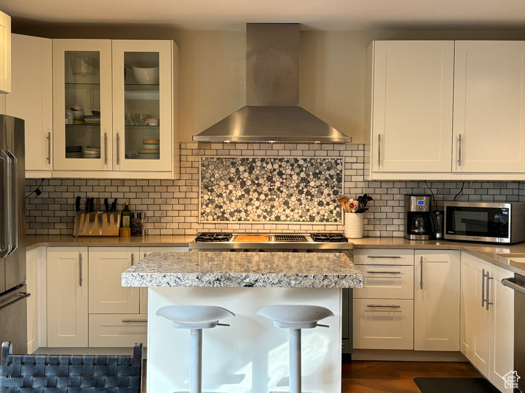 Kitchen featuring tasteful backsplash, a breakfast bar area, white cabinetry, wall chimney exhaust hood, and appliances with stainless steel finishes