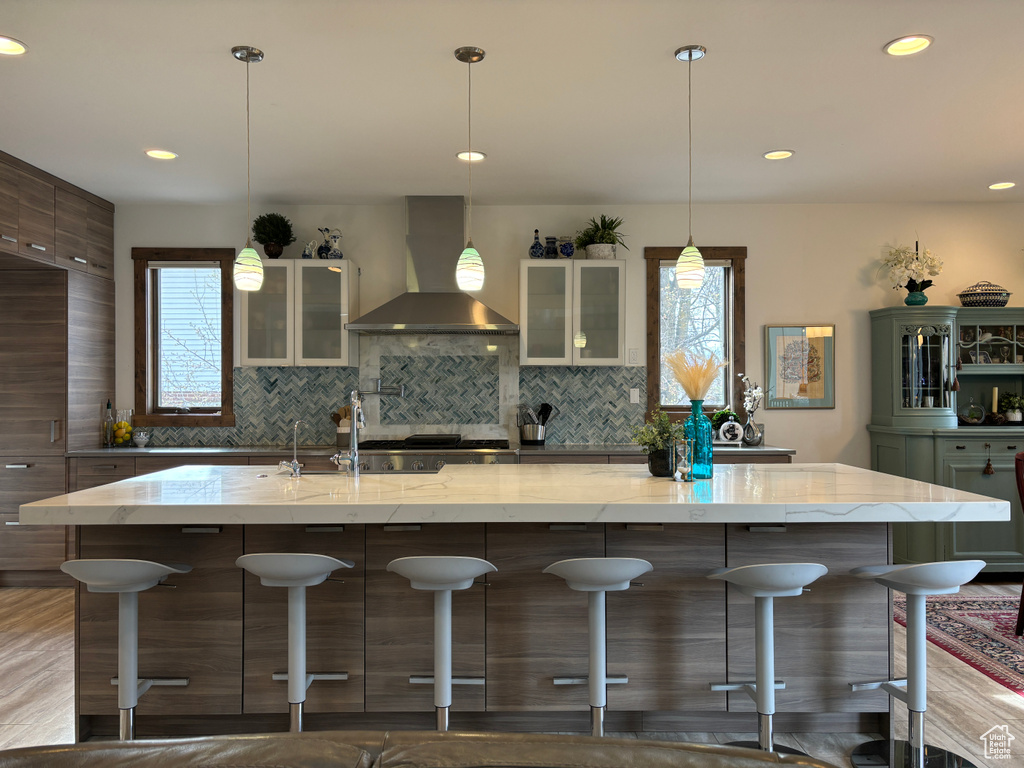 Kitchen with hanging light fixtures, a kitchen island with sink, light stone counters, wall chimney exhaust hood, and backsplash