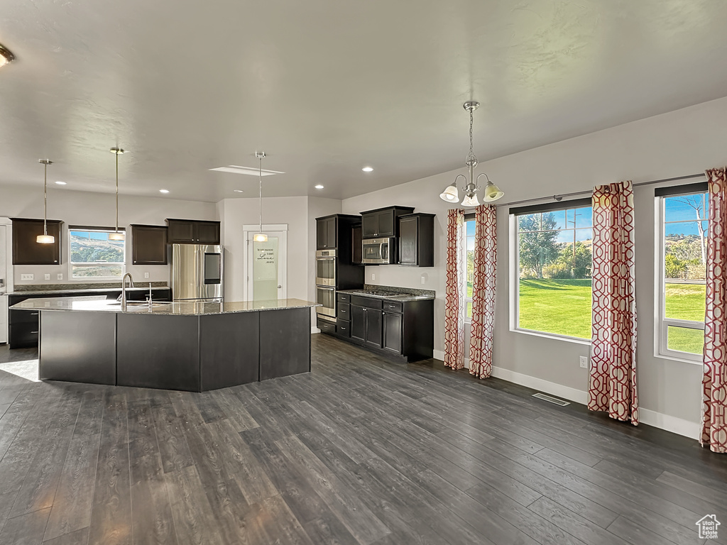 Kitchen with dark hardwood / wood-style floors, decorative light fixtures, appliances with stainless steel finishes, an island with sink, and a chandelier