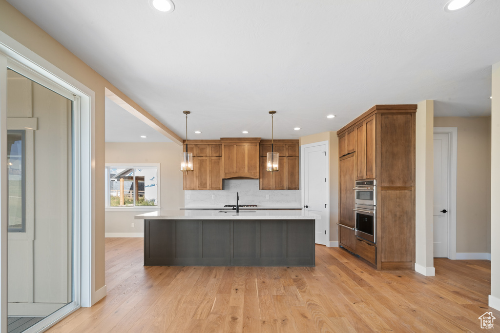 Kitchen with decorative light fixtures, stainless steel double oven, light hardwood / wood-style flooring, an island with sink, and tasteful backsplash