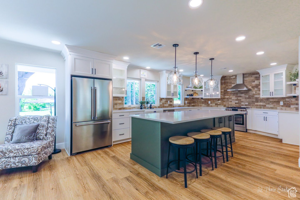 Kitchen with white cabinetry, light wood-type flooring, a kitchen island, stainless steel appliances, and wall chimney exhaust hood