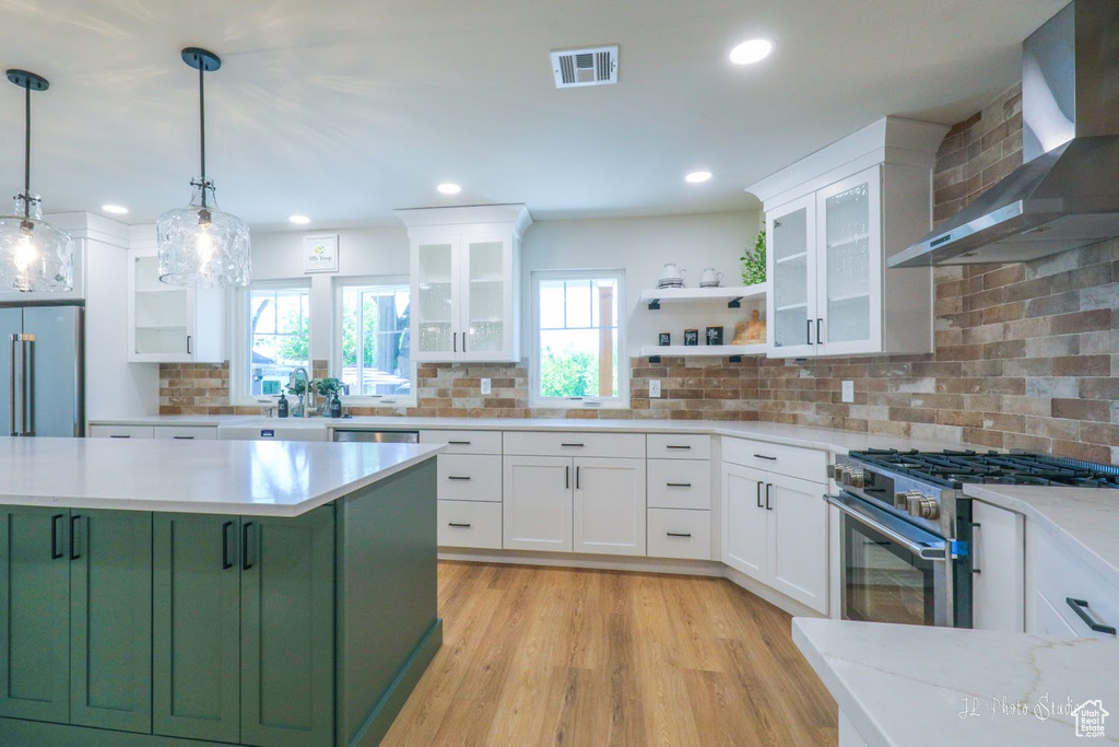 Kitchen featuring backsplash, wall chimney exhaust hood, green cabinetry, appliances with stainless steel finishes, and pendant lighting