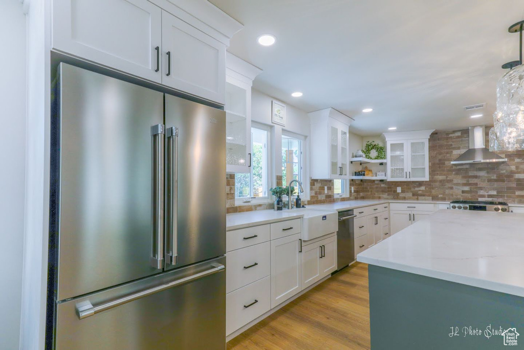 Kitchen featuring light hardwood / wood-style floors, stainless steel appliances, white cabinets, wall chimney exhaust hood, and backsplash