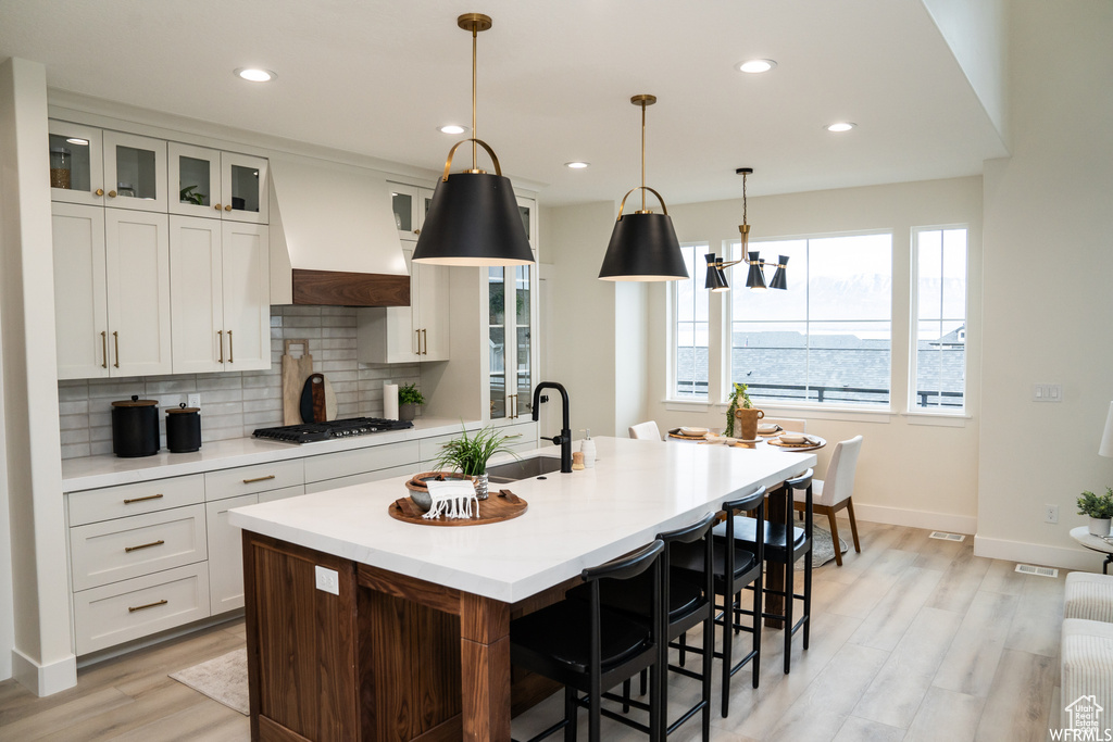 Kitchen featuring tasteful backsplash, a center island with sink, decorative light fixtures, light wood-type flooring, and custom range hood