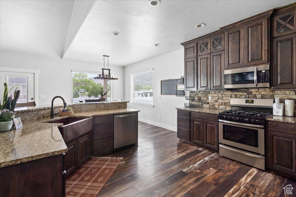 Kitchen featuring dark hardwood / wood-style floors, dark brown cabinetry, light stone counters, hanging light fixtures, and appliances with stainless steel finishes