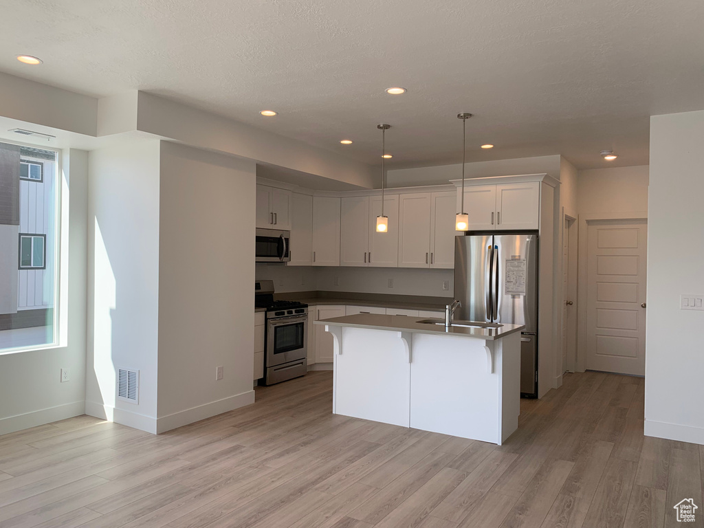 Kitchen with appliances with stainless steel finishes, white cabinetry, light wood-type flooring, and a kitchen island with sink