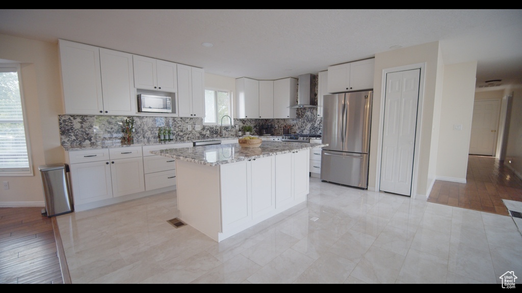 Kitchen with wall chimney range hood, white cabinetry, a kitchen island, stainless steel appliances, and backsplash