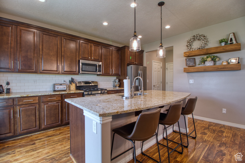 Kitchen with an island with sink, wood-type flooring, backsplash, hanging light fixtures, and appliances with stainless steel finishes