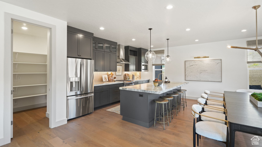 Kitchen featuring stainless steel appliances, wood-type flooring, hanging light fixtures, wall chimney exhaust hood, and a kitchen island with sink