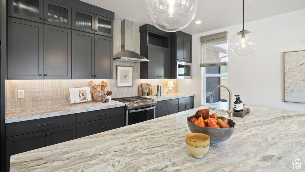 Kitchen featuring stainless steel appliances, light stone counters, wall chimney exhaust hood, decorative light fixtures, and backsplash