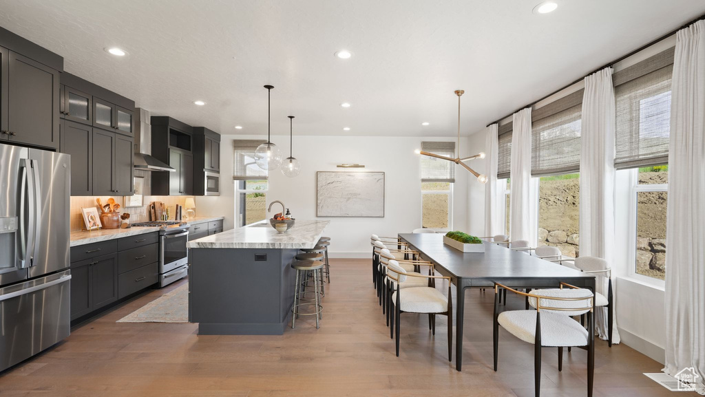 Kitchen with hanging light fixtures, a center island with sink, appliances with stainless steel finishes, wall chimney exhaust hood, and wood-type flooring